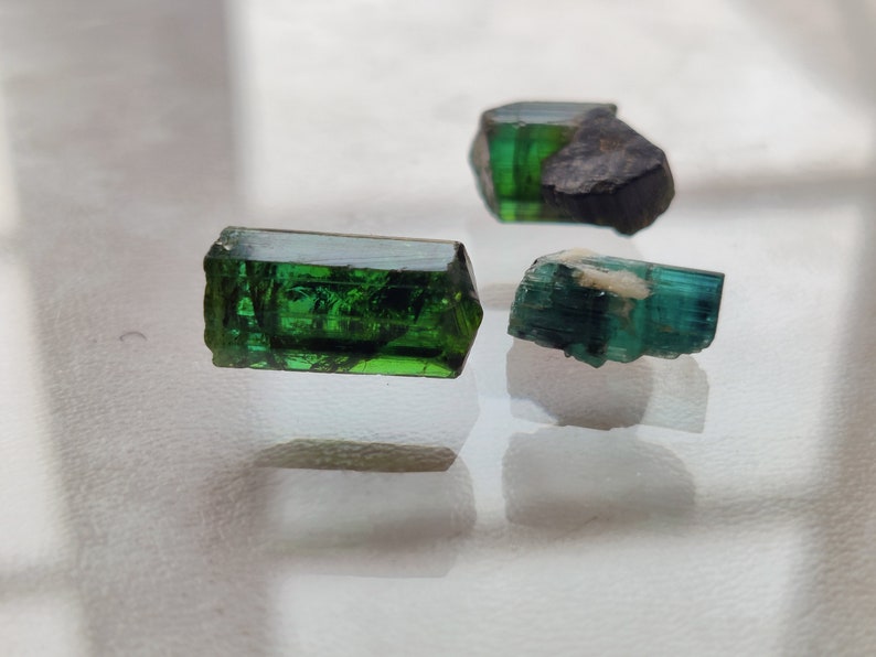 May include: Three green gemstones, two of which are long and rectangular, and one is shorter and more rounded. The gemstones are on a white surface.
