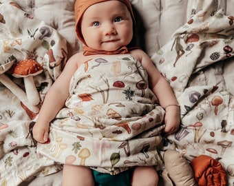 May include: A baby wearing a brown knit hat and a swaddle with a mushroom and leaf print. The baby is lying on a white quilted blanket.
