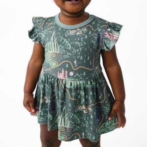May include: A green baby dress with a map-like design featuring castles, mountains, and trees. The dress has short sleeves with ruffled edges and a round neckline. The child is smiling.