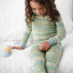 Puede incluir: Conjunto de pijama de dos piezas para ni&ntilde;os, de color verde claro, con un estampado de tem&aacute;tica de Pascua. El pijama presenta una parte superior de manga larga y pantalones a juego con un dise&ntilde;o repetido de huevos, pollitos y conejitos. Los pu&ntilde;os son de color azul claro.