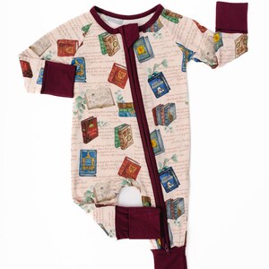 May include: A light pink baby sleeper with a repeating pattern of colorful books and handwritten script. The sleeper has a burgundy zipper, trim, and cuffs. The footed sleeper is designed for infants and toddlers.