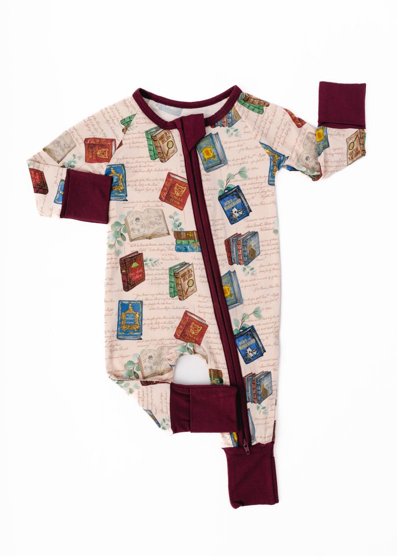 May include: A light pink baby sleeper with a repeating pattern of colorful books and handwritten script. The sleeper has a burgundy zipper, trim, and cuffs. The footed sleeper is designed for infants and toddlers.