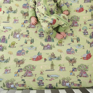 May include: A light green fitted cot sheet with a whimsical town map design. The sheet features illustrations of buildings, trees, and people. A baby in a matching green onesie is lying on the sheet.