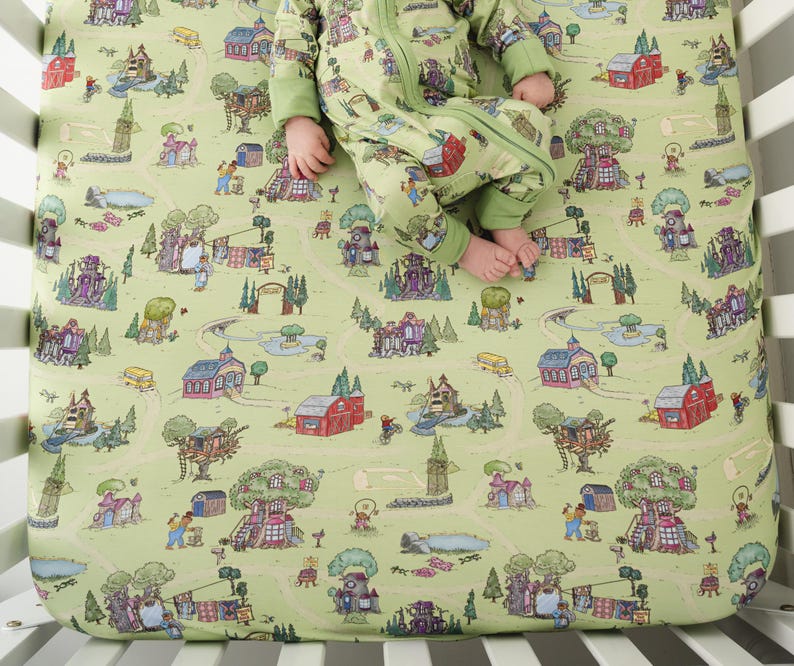 May include: A light green fitted cot sheet with a whimsical town map design. The sheet features illustrations of buildings, trees, and people. A baby in a matching green onesie is lying on the sheet.