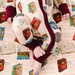 May include: A beige baby sleeper with a repeating pattern of colorful books, including titles like "Sherlock Holmes" and "The Jungle Book." The sleeper has burgundy cuffs and a burgundy zipper. The background is a light pink with script.