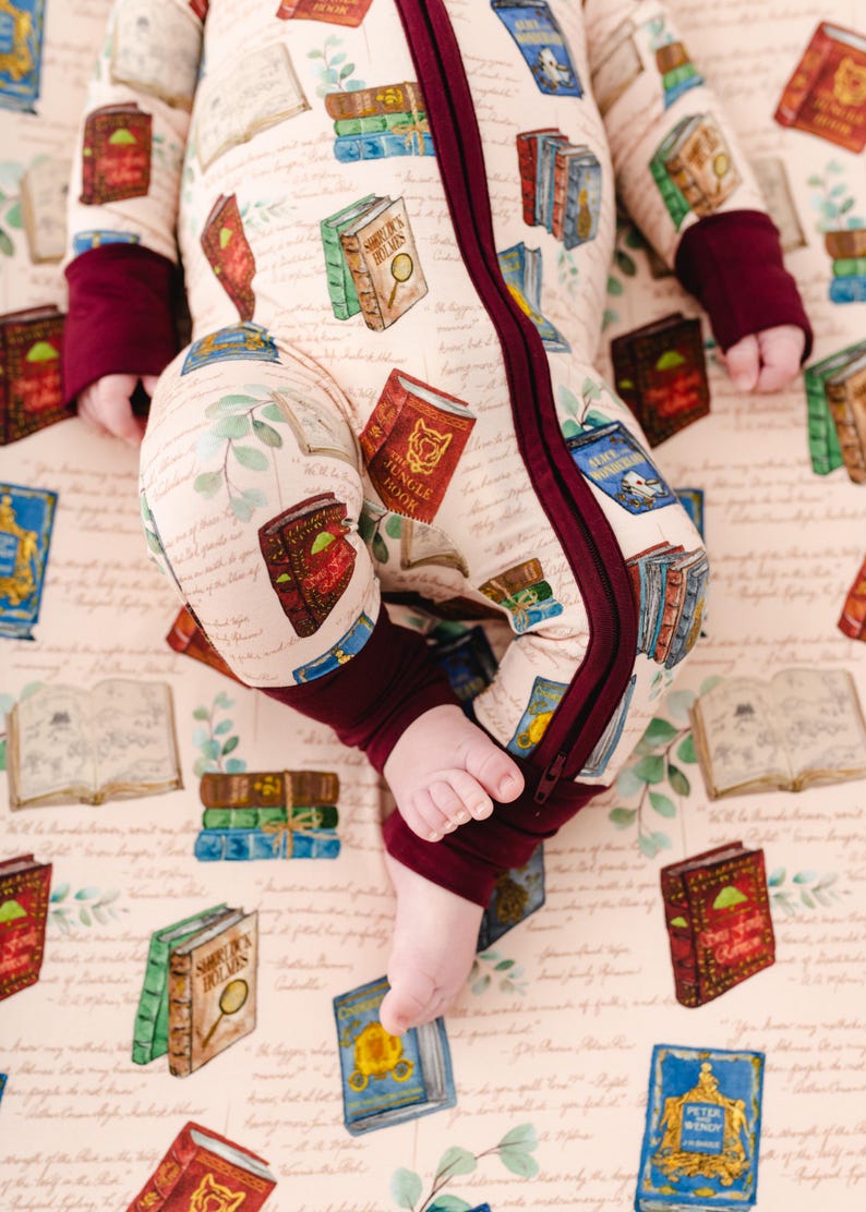 May include: A beige baby sleeper with a repeating pattern of colorful books, including titles like "Sherlock Holmes" and "The Jungle Book." The sleeper has burgundy cuffs and a burgundy zipper. The background is a light pink with script.