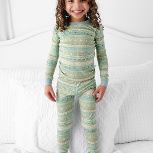 Puede incluir: Un ni&ntilde;o lleva un pijama verde menta con un patr&oacute;n repetido de pollitos amarillos, zanahorias y otros dise&ntilde;os. La parte superior de manga larga y los pantalones tienen pu&ntilde;os de color azul claro. El ni&ntilde;o est&aacute; de pie sobre una cama blanca.