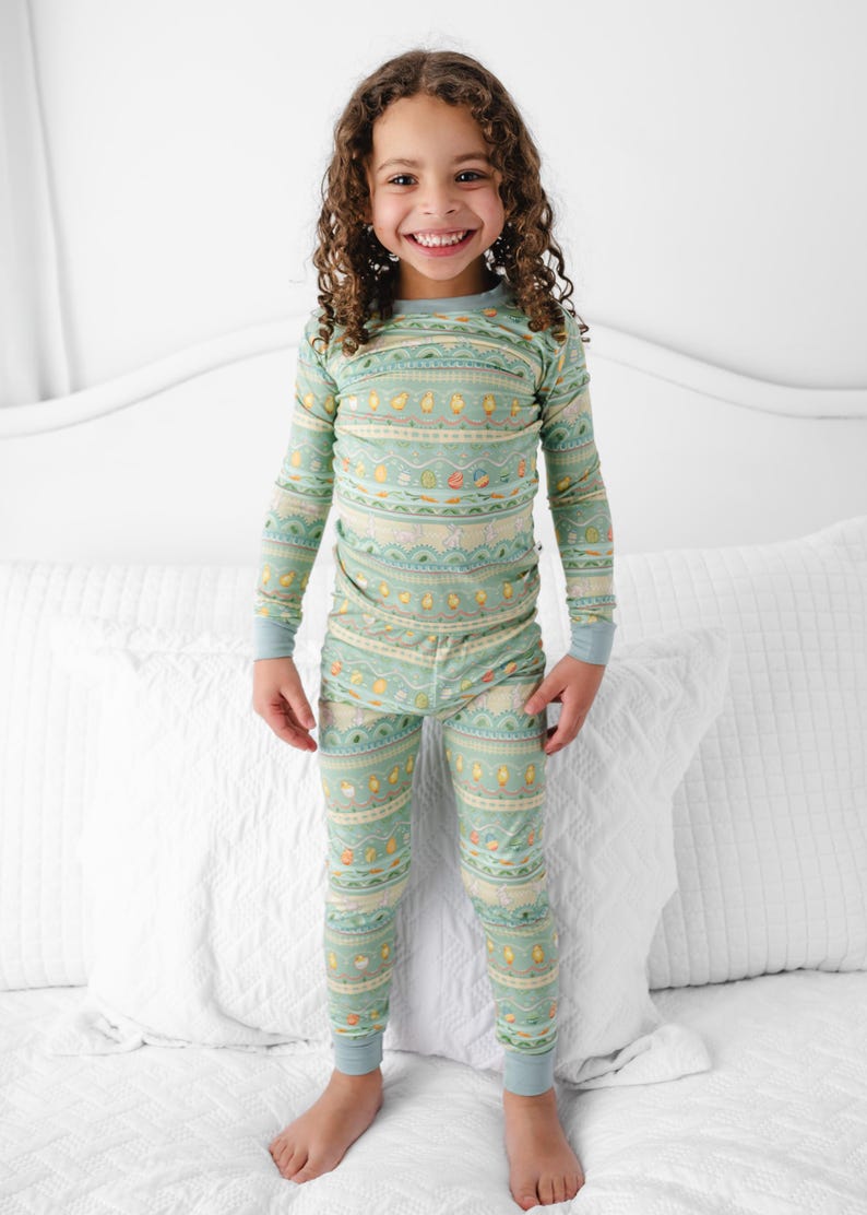 Puede incluir: Un ni&ntilde;o lleva un pijama verde menta con un patr&oacute;n repetido de pollitos amarillos, zanahorias y otros dise&ntilde;os. La parte superior de manga larga y los pantalones tienen pu&ntilde;os de color azul claro. El ni&ntilde;o est&aacute; de pie sobre una cama blanca.