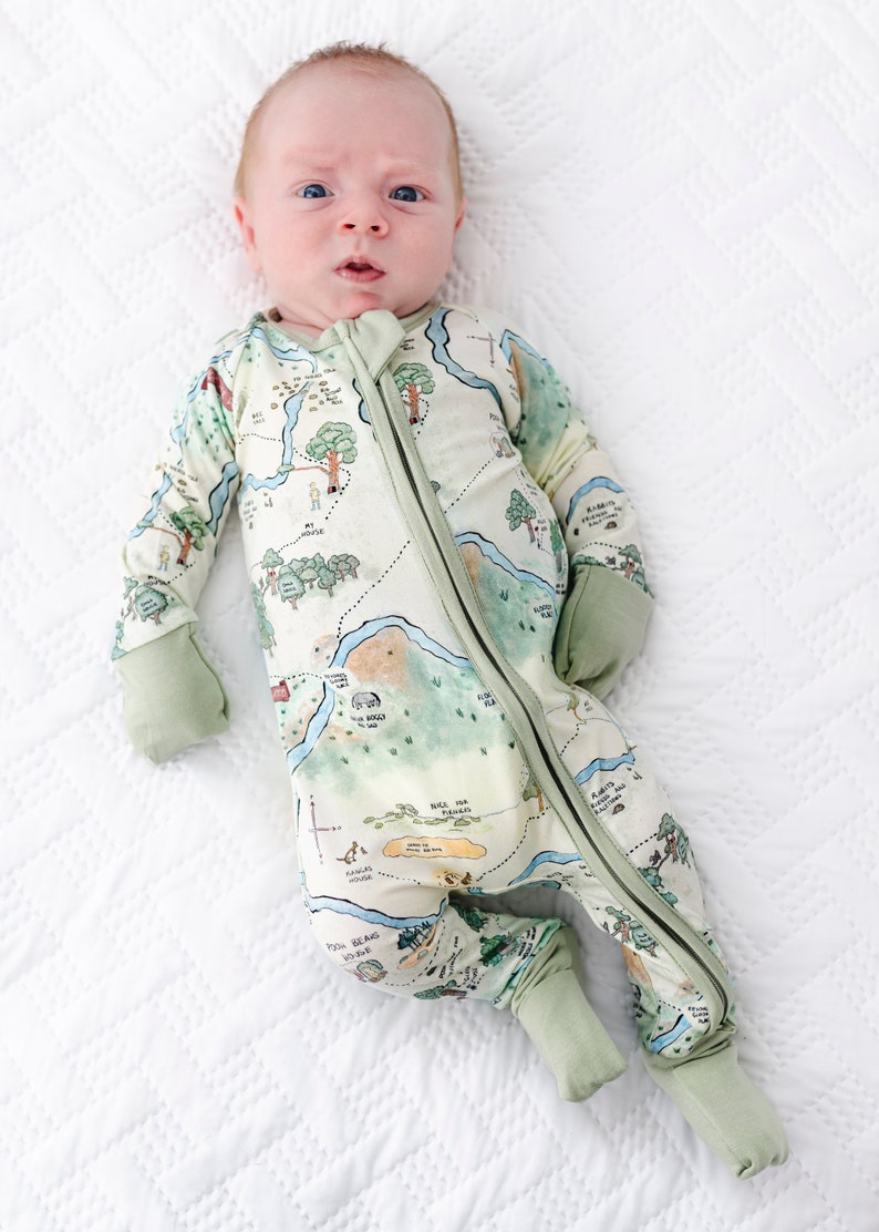 May include: A baby wearing a green and white long-sleeved onesie with a map design. The onesie has a zipper down the front and the map design includes a variety of animals and landmarks.