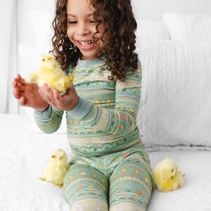 Puede incluir: Un ni&ntilde;o con un pijama verde menta con un estampado de Pascua, sosteniendo un pollito amarillo. El pijama presenta conejitos, huevos y otros motivos primaverales. Dos pollitos adicionales est&aacute;n en la colcha blanca.
