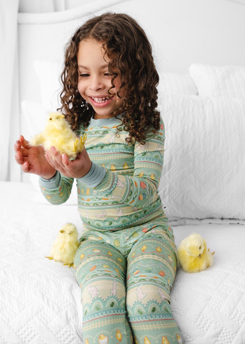 Puede incluir: Un ni&ntilde;o con un pijama verde menta con un estampado de Pascua, sosteniendo un pollito amarillo. El pijama presenta conejitos, huevos y otros motivos primaverales. Dos pollitos adicionales est&aacute;n en la colcha blanca.