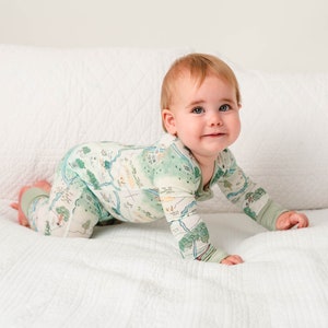 May include: A baby in a green and white patterned long-sleeved onesie crawls on a white bed. The onesie has a map-like design with trees, rivers, and other natural features.
