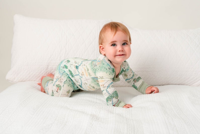 May include: A baby in a green and white patterned long-sleeved onesie crawls on a white bed. The onesie has a map-like design with trees, rivers, and other natural features.