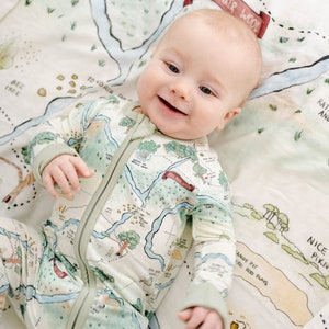 May include: A baby wearing a green and white long-sleeved onesie with a map print. The onesie has a zipper down the front. The baby is lying on a white blanket with a map print. The map print includes the text "100 Aker Wood".