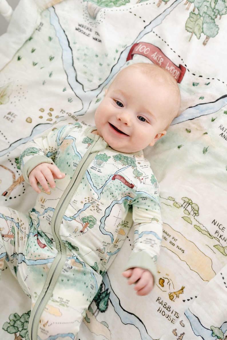 May include: A baby wearing a green and white long-sleeved onesie with a map print. The onesie has a zipper down the front. The baby is lying on a white blanket with a map print. The map print includes the text "100 Aker Wood".