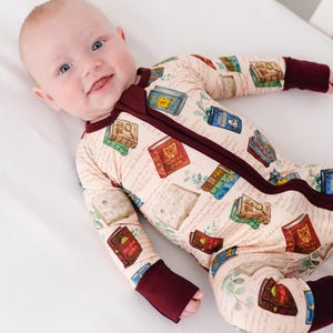 May include: A baby wearing a beige footed pajama with a book-themed print. The pajama features a burgundy zipper, cuffs, and neckline. The print includes various book illustrations and handwritten script.