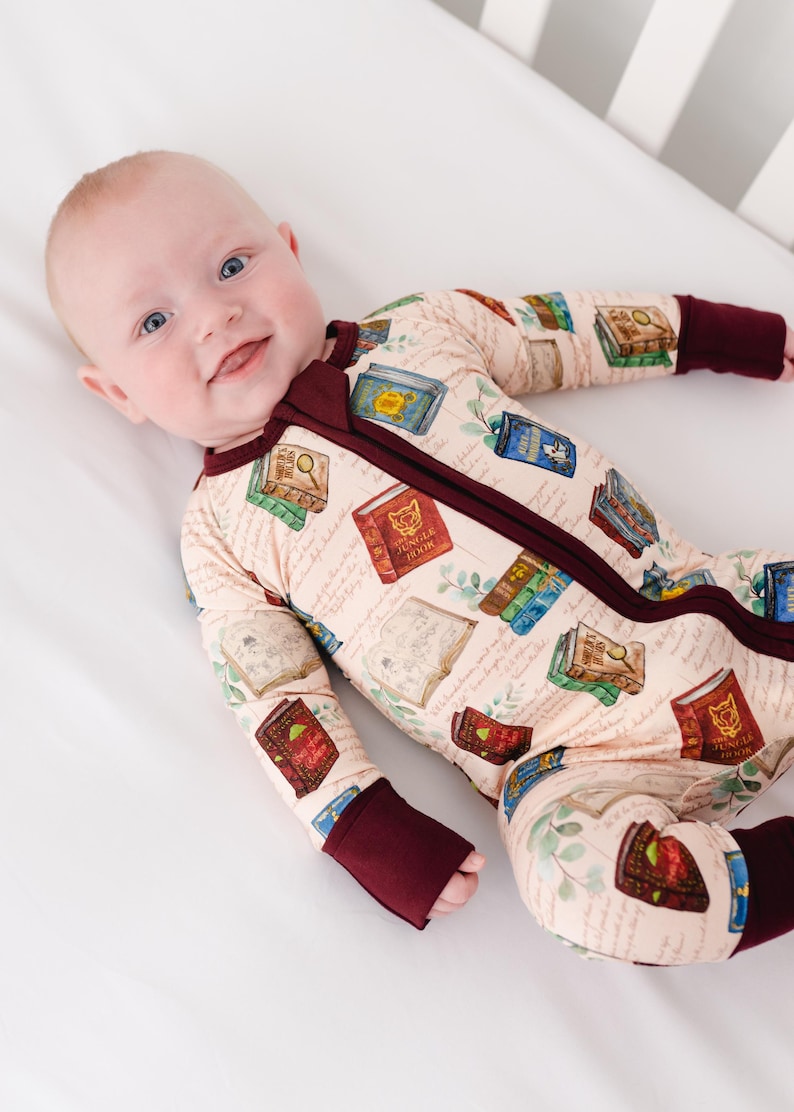 May include: A baby wearing a beige footed pajama with a book-themed print. The pajama features a burgundy zipper, cuffs, and neckline. The print includes various book illustrations and handwritten script.