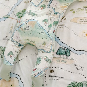 May include: A white baby onesie with a map of the Hundred Acre Wood from Winnie the Pooh. The onesie has green, blue, and brown details and features the text "Pooh Trap for Heffalumps", "Piglets House", "Six Pine Trees", and "Sandy Pit Where Roo", among other locations.