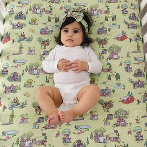 May include: A baby lies on a cot sheet with a whimsical town map design. The sheet features illustrations of houses, trees, and roads in shades of green, red, and purple. The baby wears a white onesie and a matching headband.