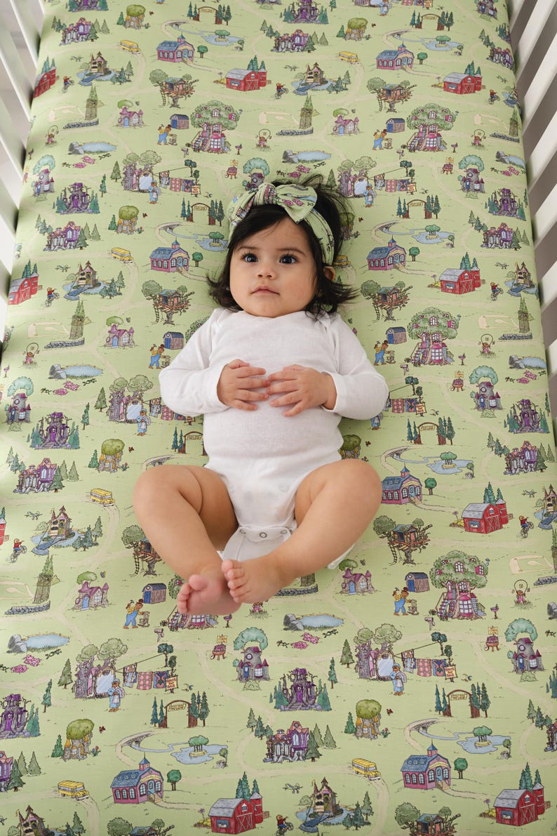 May include: A baby lies on a cot sheet with a whimsical town map design. The sheet features illustrations of houses, trees, and roads in shades of green, red, and purple. The baby wears a white onesie and a matching headband.