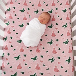 Könnte beinhalten: Ein Baby schläft in einer weißen Decke gewickelt auf einem rosa Bettlaken. Das Laken ist mit Illustrationen von grünen Hexen, rosa Kleidern, schwarzen Hexenhüten, Kronen und Zauberbüchern gemustert.