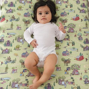 May include: A baby in a white babygrow lies on a cot sheet with a whimsical town map design. The sheet features illustrations of houses, trees, and roads in shades of green, red, and purple. The baby has dark hair and is looking at the camera.