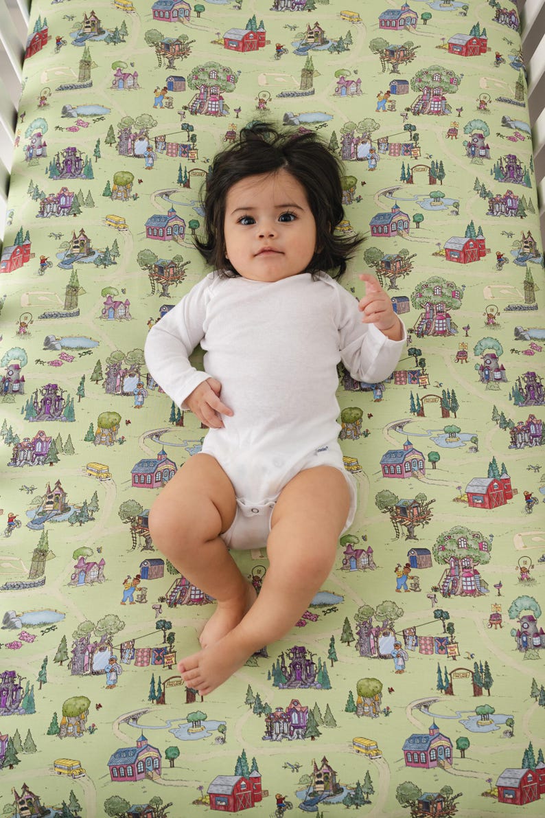 May include: A baby in a white babygrow lies on a cot sheet with a whimsical town map design. The sheet features illustrations of houses, trees, and roads in shades of green, red, and purple. The baby has dark hair and is looking at the camera.