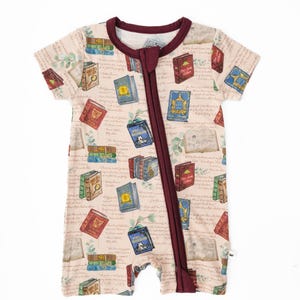 May include: A short-sleeved, one-piece baby romper with a cream background and a repeating pattern of colorful books. The romper has a burgundy zipper and trim. The fabric is covered in script and botanical illustrations.