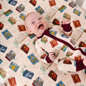 May include: A baby wearing a cream-colored onesie with a burgundy zipper and trim, lying on a crib sheet. The sheet and onesie are patterned with illustrations of colorful books. The background is a light beige with script and botanical accents.