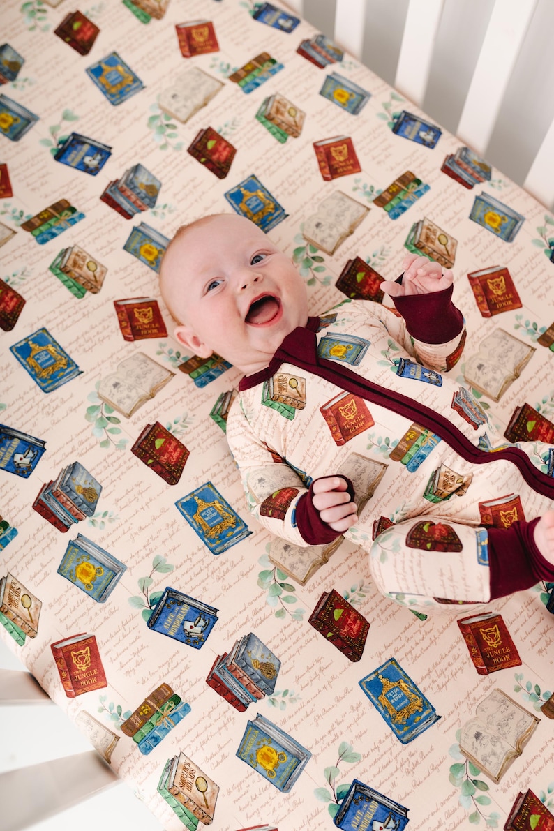 May include: A baby wearing a cream-colored onesie with a burgundy zipper and trim, lying on a crib sheet. The sheet and onesie are patterned with illustrations of colorful books. The background is a light beige with script and botanical accents.