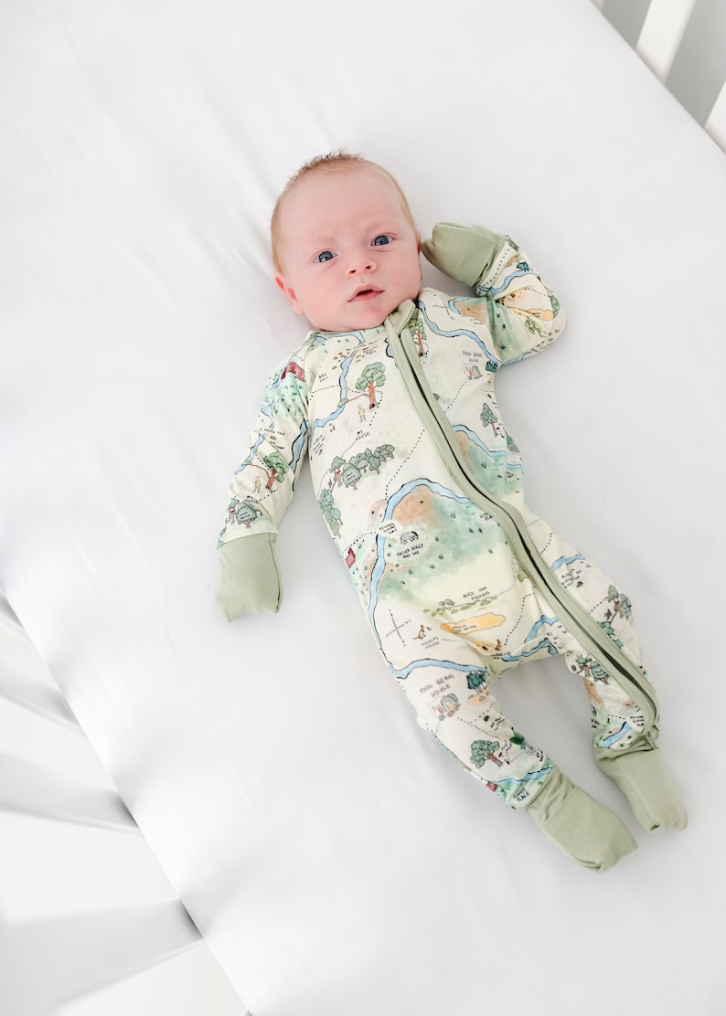 May include: A baby wearing a green and white map-print footed pajamas with a zipper closure. The pajamas have green cuffs on the sleeves and legs.