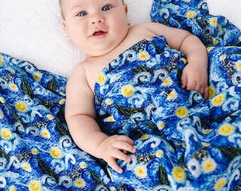 May include: A baby wrapped in a blue blanket with a swirling, starry night design. The blanket features yellow and white accents, reminiscent of Van Gogh's "Starry Night" painting. The baby is lying on a white surface.