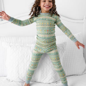 Puede incluir: Un conjunto de pijama de dos piezas para ni&ntilde;os en color verde claro con un patr&oacute;n repetido de varias formas y colores. La parte superior de manga larga y los pantalones tienen pu&ntilde;os azules. El ni&ntilde;o sonr&iacute;e y salta sobre una cama blanca.