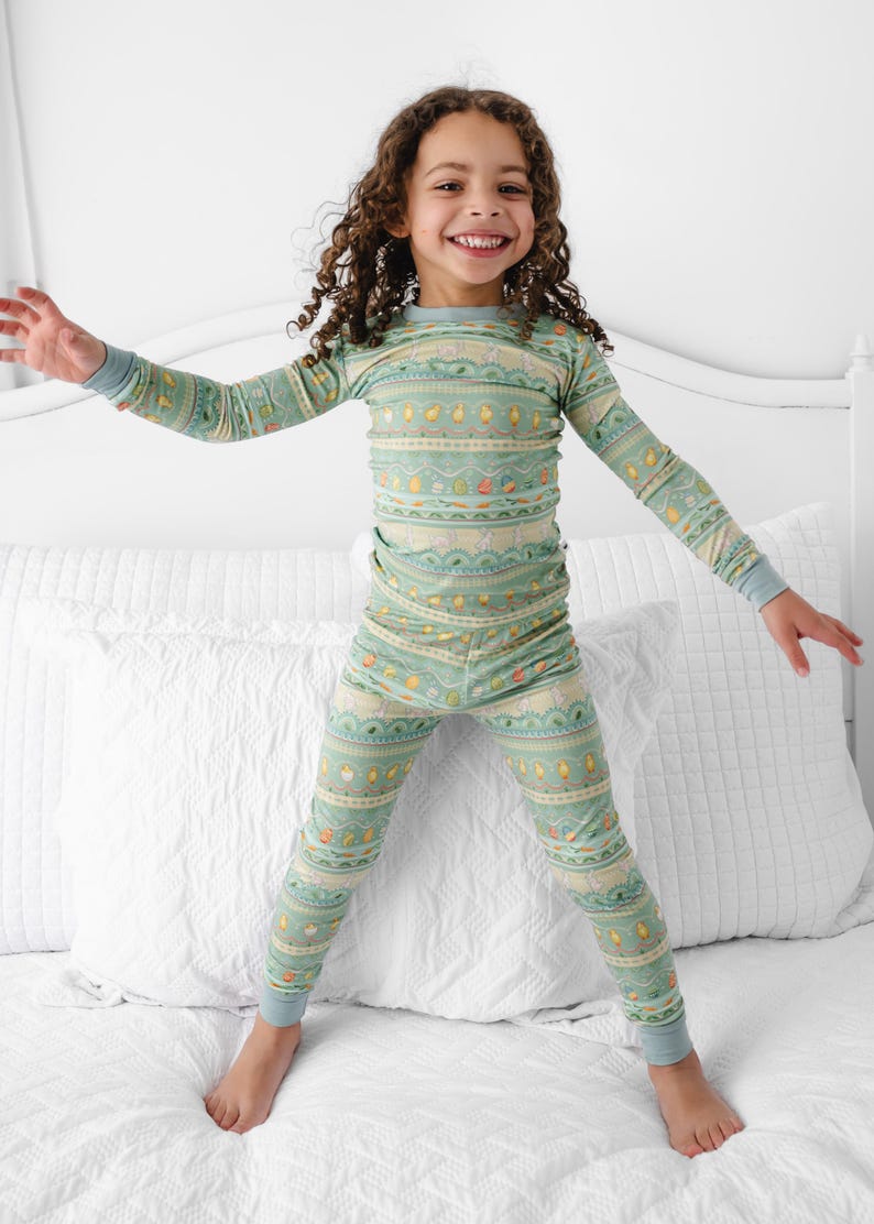 Puede incluir: Un conjunto de pijama de dos piezas para ni&ntilde;os en color verde claro con un patr&oacute;n repetido de varias formas y colores. La parte superior de manga larga y los pantalones tienen pu&ntilde;os azules. El ni&ntilde;o sonr&iacute;e y salta sobre una cama blanca.