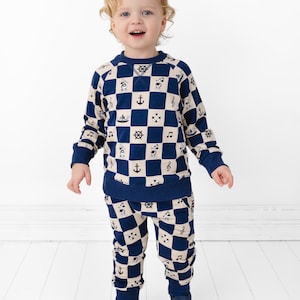 May include: A toddler wearing a navy blue and cream-colored nautical-themed outfit. The two-piece set features a checkered pattern with anchors, musical notes, and ship wheels. The outfit includes a long-sleeved top and matching pants.