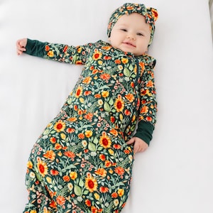 Pode incluir: Um bebê vestindo um saco de dormir verde e amarelo com estampa floral e uma faixa de cabeça combinando. O saco de dormir tem um zíper e a faixa de cabeça está amarrada em um laço.