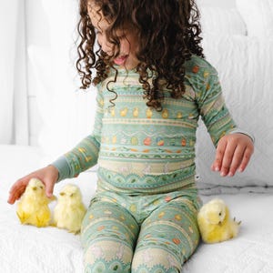 Puede incluir: Un conjunto de pijama de dos piezas de color verde claro para ni&ntilde;os, con un estampado de tem&aacute;tica de Pascua. El estampado incluye huevos, pollitos y conejos. El ni&ntilde;o est&aacute; sentado en una cama blanca con tres pollitos amarillos.