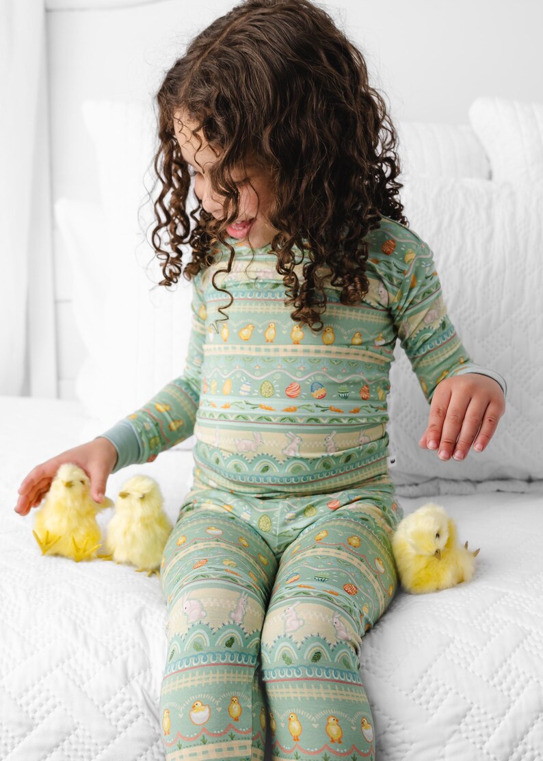 Puede incluir: Un conjunto de pijama de dos piezas de color verde claro para ni&ntilde;os, con un estampado de tem&aacute;tica de Pascua. El estampado incluye huevos, pollitos y conejos. El ni&ntilde;o est&aacute; sentado en una cama blanca con tres pollitos amarillos.