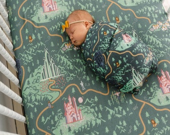 May include: A baby swaddled in a green and yellow fabric with a map design featuring a yellow brick road, a castle, and a green emerald city. The fabric is printed with a whimsical design of a magical land.