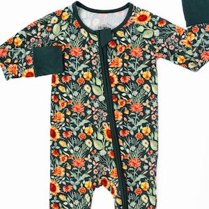 Pode incluir: Um macacão verde de manga comprida com zíper, com estampa floral de girassóis, flores laranja, amarelas e vermelhas. O macacão tem um zíper verde e punhos verdes.