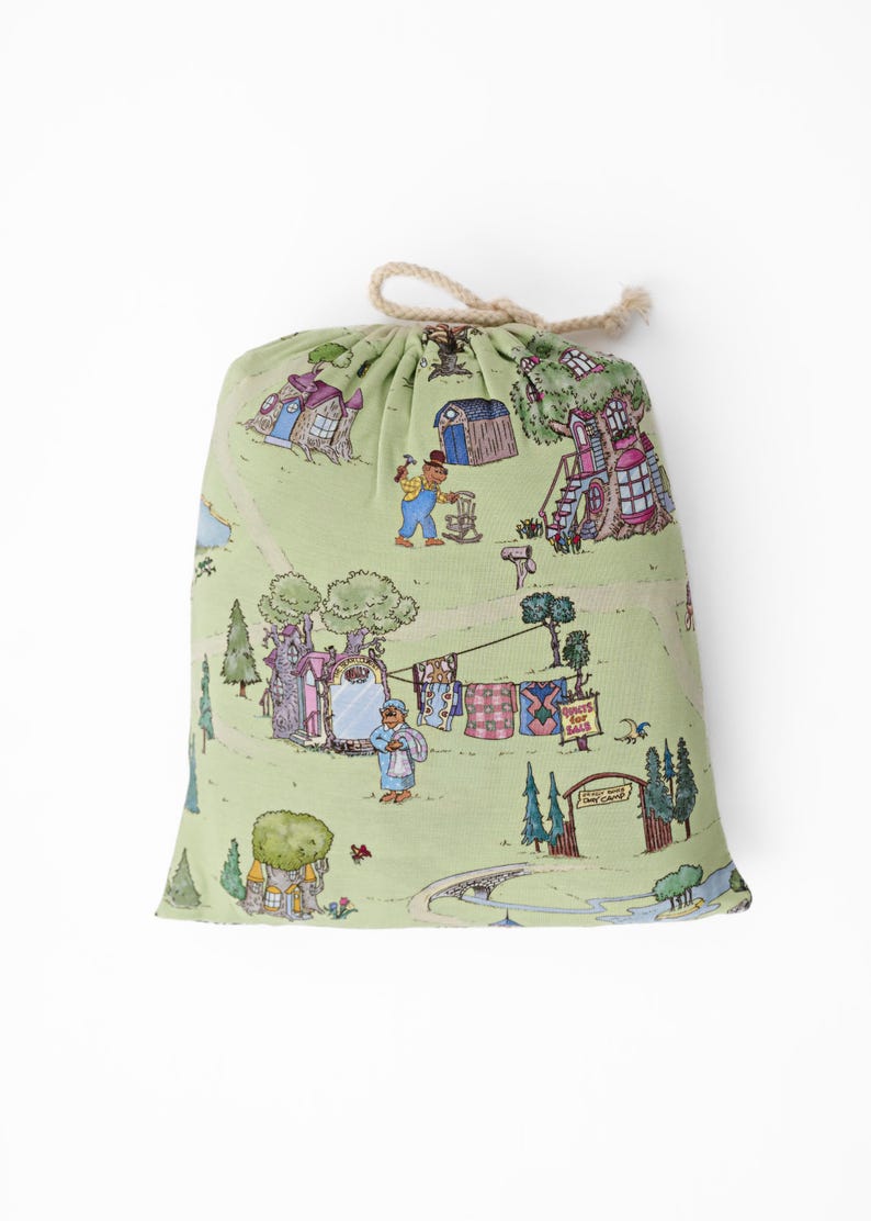 May include: A drawstring bag featuring a light green illustrated pattern of a village scene with houses, trees, and figures. The bag is tied with a natural rope. The design has a whimsical, storybook aesthetic, with a charming, playful feel.