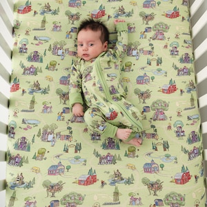 May include: A baby in a green zip-up sleepsuit lies on a cot sheet with a whimsical town scene print. The sheet and sleepsuit are a light green colour. The town scene includes houses, trees, and roads.