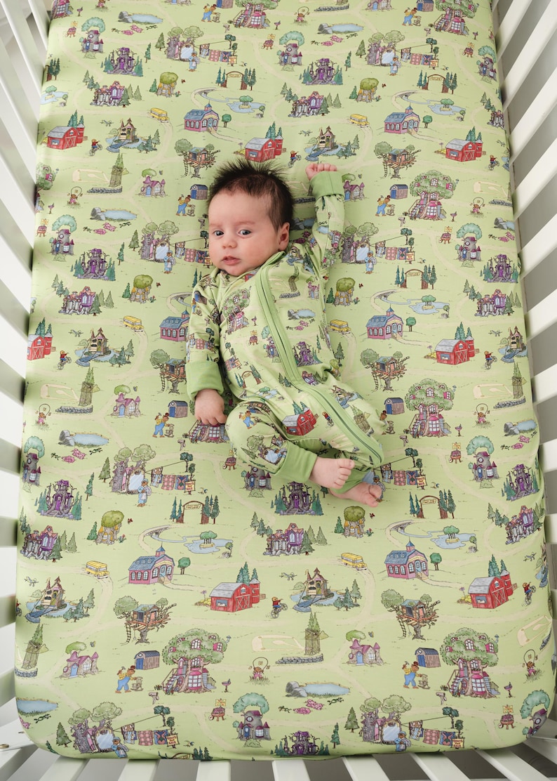 May include: A baby in a green zip-up sleepsuit lies on a cot sheet with a whimsical town scene print. The sheet and sleepsuit are a light green colour. The town scene includes houses, trees, and roads.