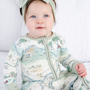 May include: A baby wearing a green and white long-sleeved onesie with a map print. The onesie has a zipper down the front and fold-over cuffs. The baby is wearing a green headband with a bow on top.