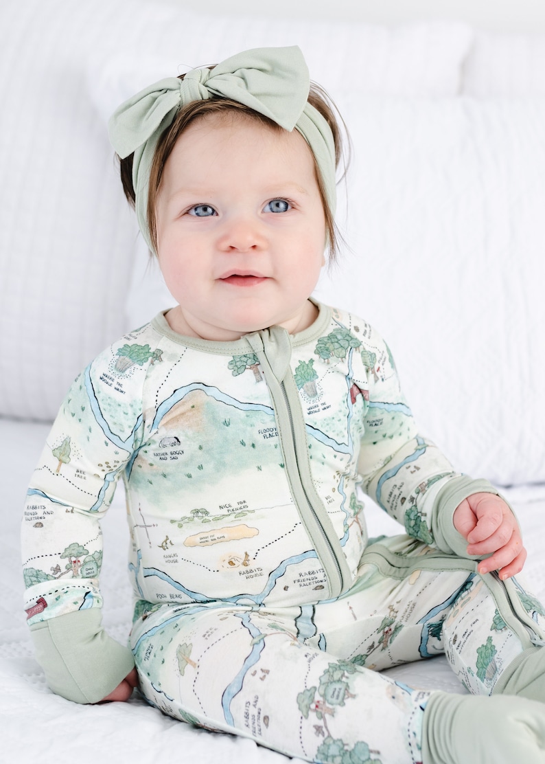 May include: A baby wearing a green and white long-sleeved onesie with a map print. The onesie has a zipper down the front and fold-over cuffs. The baby is wearing a green headband with a bow on top.