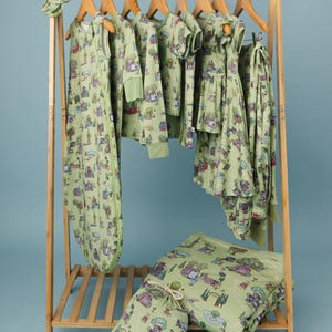 May include: A selection of light green baby clothes and accessories, featuring a village print. The items include a sleep sack, shirts, trousers, and a headband, displayed on a wooden clothes rack, with a matching blanket and storage bag.
