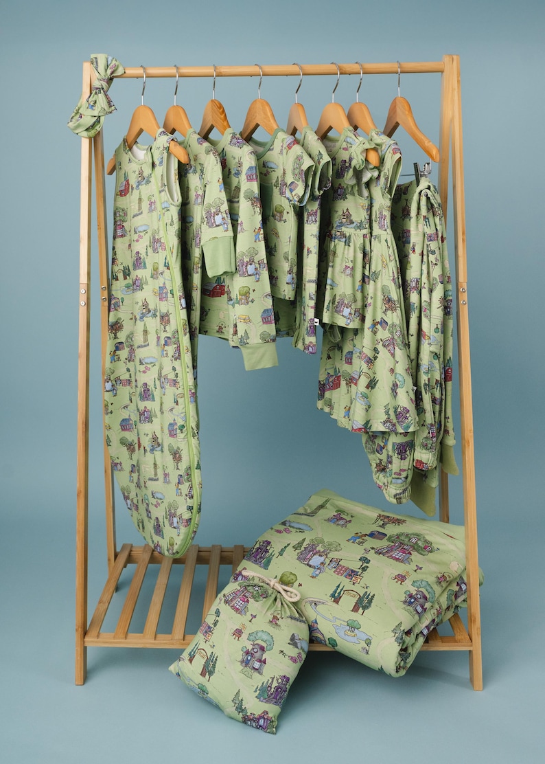 May include: A selection of light green baby clothes and accessories, featuring a village print. The items include a sleep sack, shirts, trousers, and a headband, displayed on a wooden clothes rack, with a matching blanket and storage bag.
