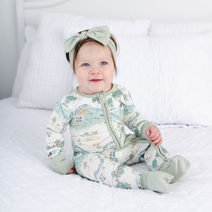 May include: A baby wearing a green and white map print footed pajamas with a green bow headband. The baby is sitting on a white bed.