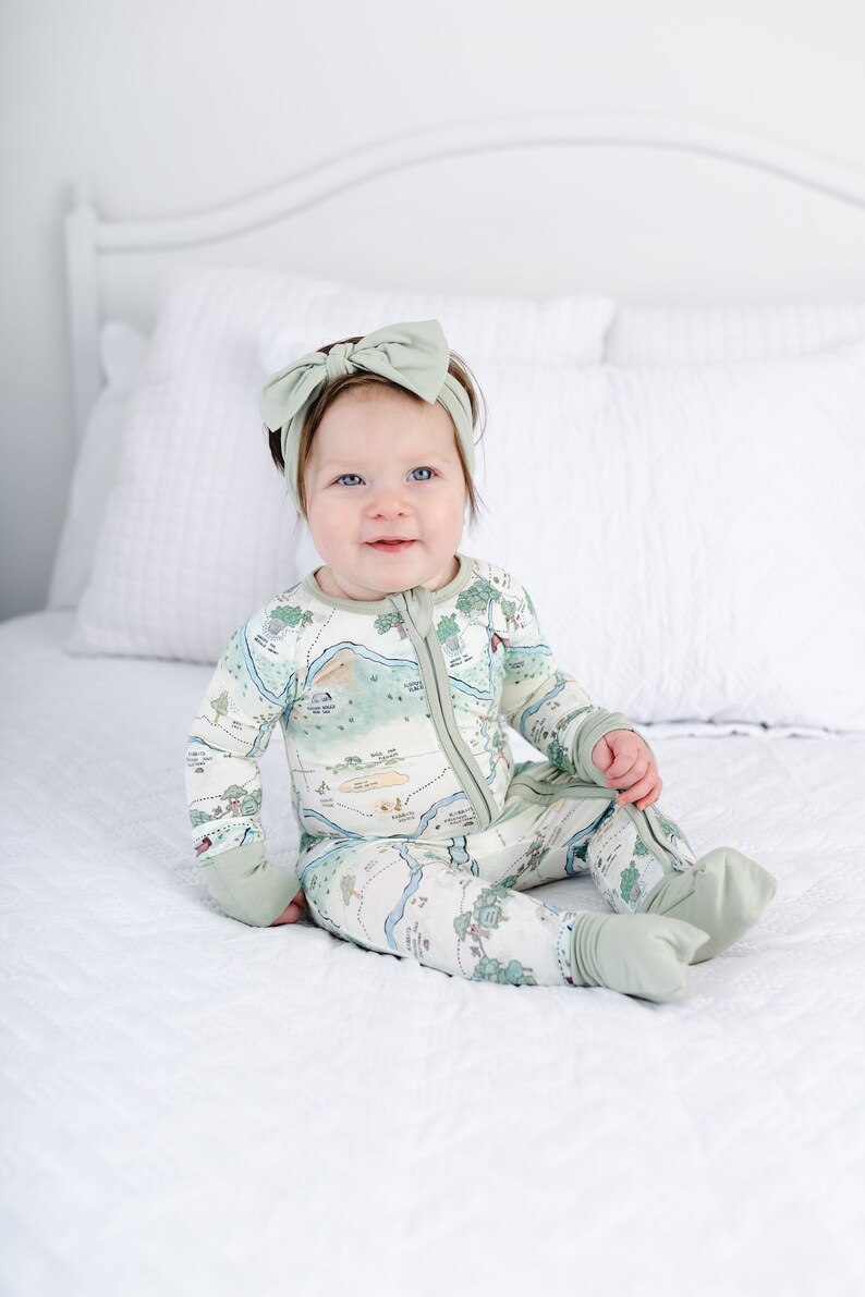 May include: A baby wearing a green and white map print footed pajamas with a green bow headband. The baby is sitting on a white bed.