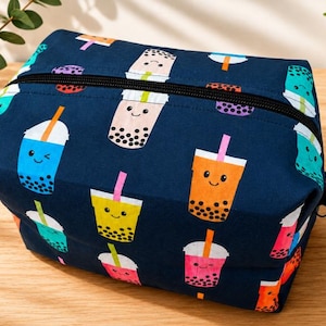 May include: A navy blue rectangular zippered pouch featuring a repeating pattern of colorful bubble tea cups with smiling faces. Each cup has a straw and black tapioca pearls at the bottom. The pouch has a black zipper closure and a small side tab.
